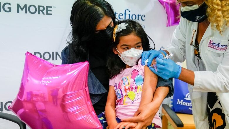 A young masked girl holding a bright pink star-shaped balloon sits on her mother's lap as a nurse gives her a shot in the arm.