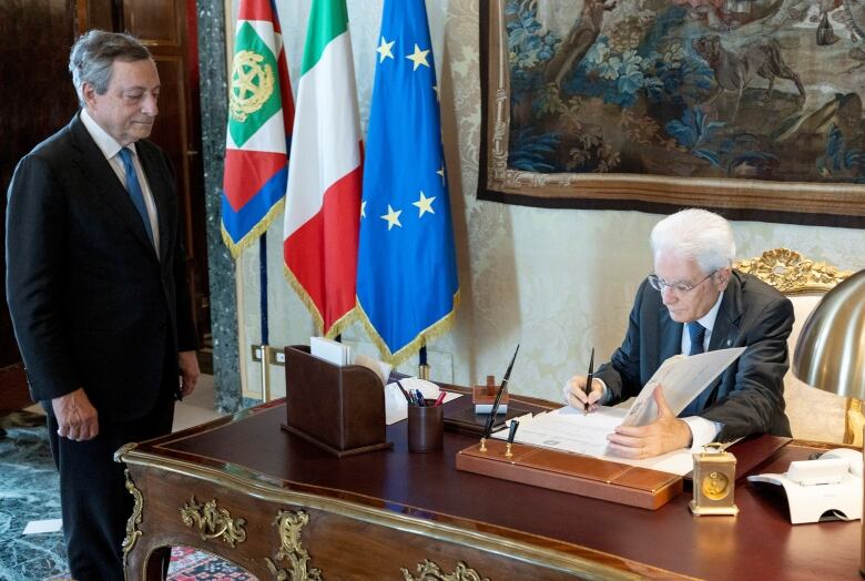 A man stands near a desk, where an older man signs an open book.