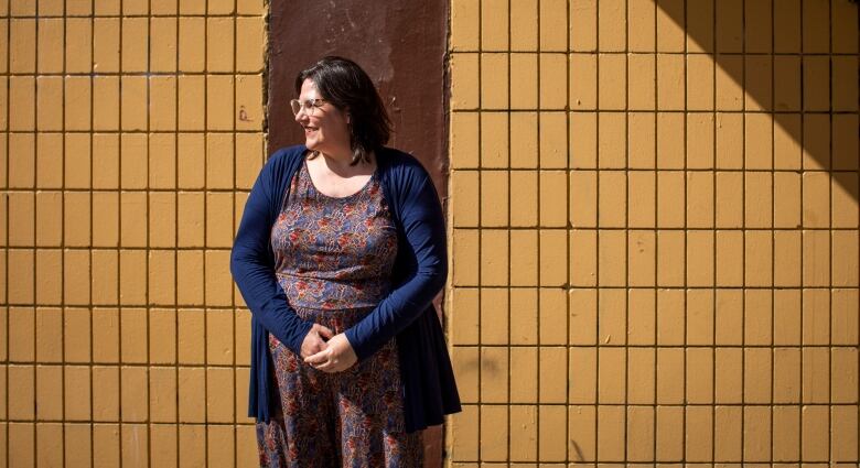 Carmen Lansdowne stands against a tiled wall. She is a wearing a blue dress with red designs, along with a blue long-sleeved blouse.