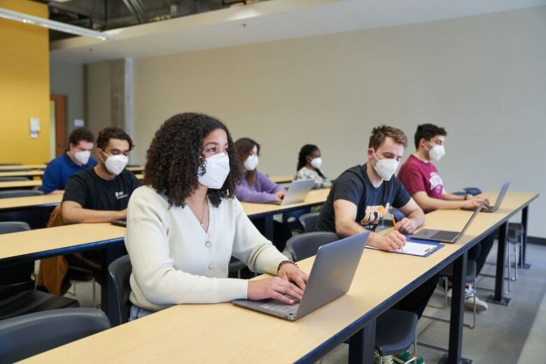 Students sit behind computers in a classroom, and all are wearing KN95 masks.