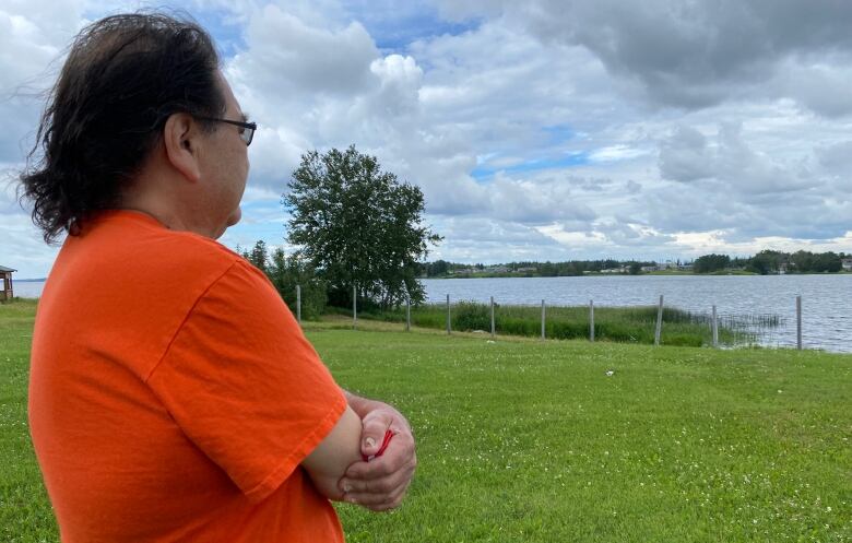 A man in an orange shirt stands looking out over a green field and a nearby river.