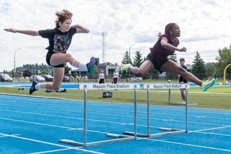 Two female athletes jump over hurdles.