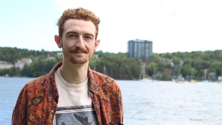 A person stands next to the water with trees, boats and buildings in the background.