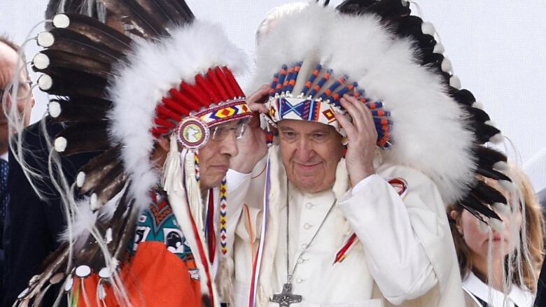 Two men (a Chief and the Pope) facing each other, each wearing a traditional headdress. 