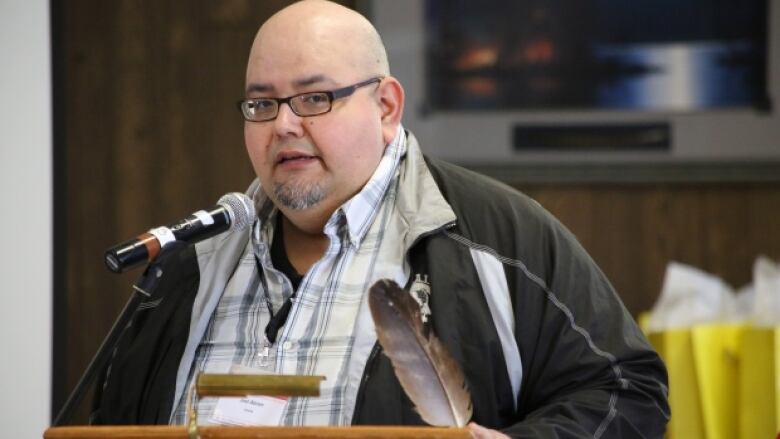 A First Nations man holds a feather and speaks into a microphone at a podium. 