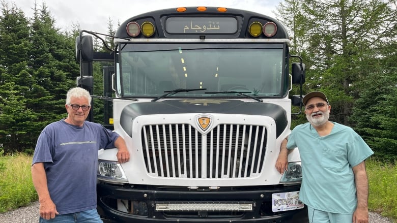 Two middle-aged men lean against the hood of a bus; one on the left and the other on the right. Both smile into the camera.