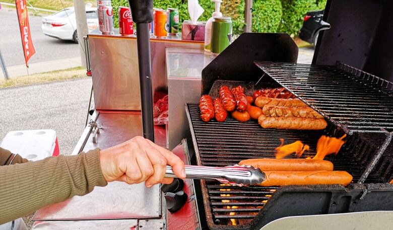 A hand holds tongs to move a hot dog on a barbecue with flames.