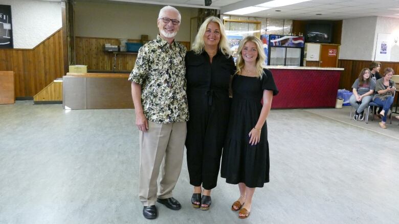 An older man, a woman and a younger woman stands side-by-side in a community centre space, smiling.