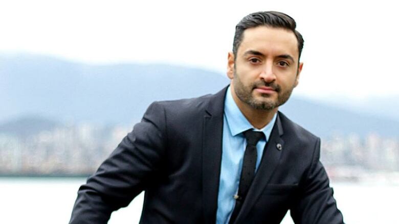 A man with olive skin, slicked black hair and a light salt-and-pepper beard poses in a dark suit with light blue shirt in front of an out-of-focus background showing Burrard Inlet and the North Shore.