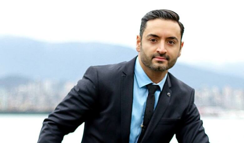 A man with olive skin, slicked black hair and a light salt-and-pepper beard poses in a dark suit with light blue shirt in front of an out-of-focus background showing Burrard Inlet and the North Shore.