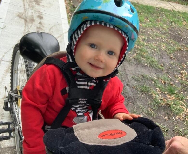 A toddler wearing a bike helmet sits on the back of a bike in a front yard.