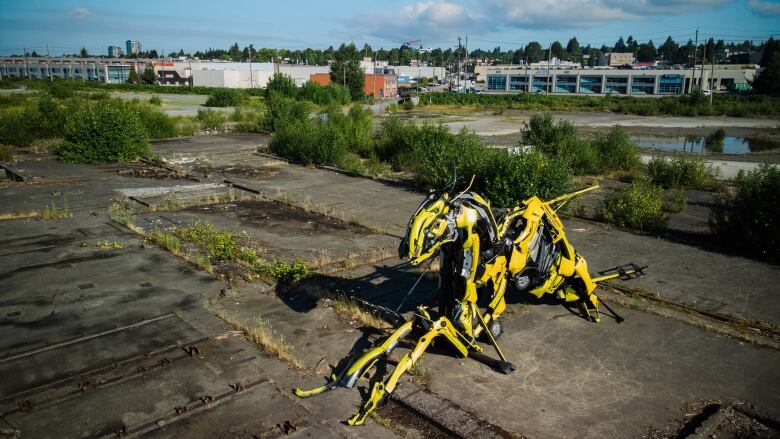 A giant yellow bee-like statue sits on an empty industrial piece of land, with industrial buildings visible in the background.