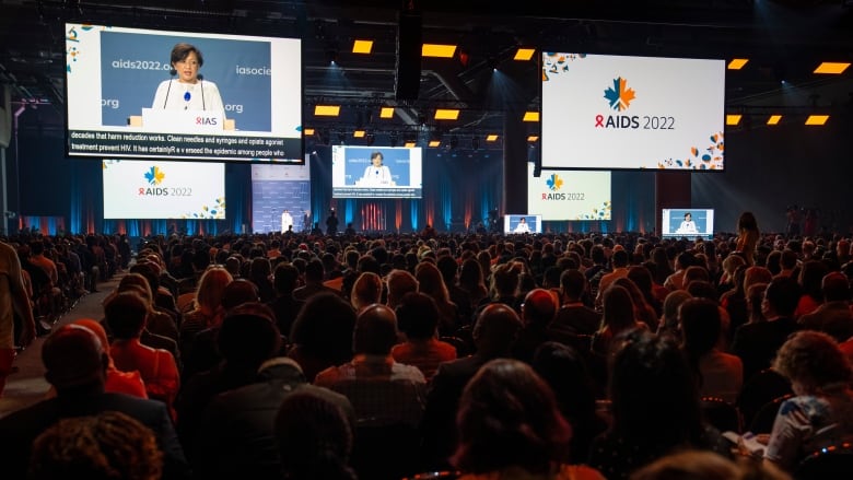 Dark conference room full of people, woman on big screen speaks to them