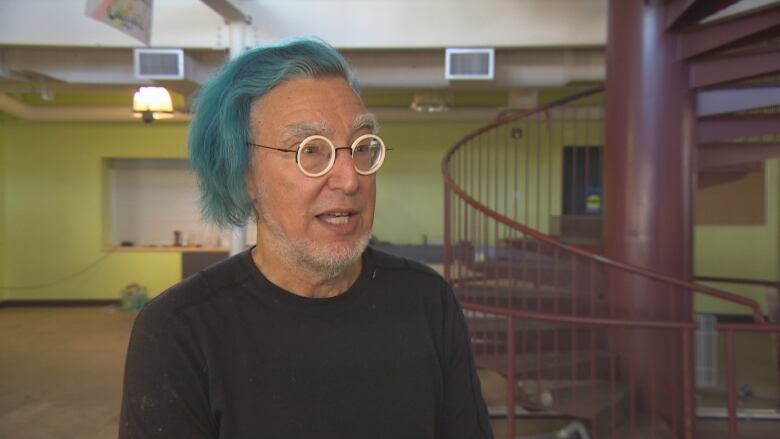 A man with round glasses and blue hair speaks in front of a spiral staircase.