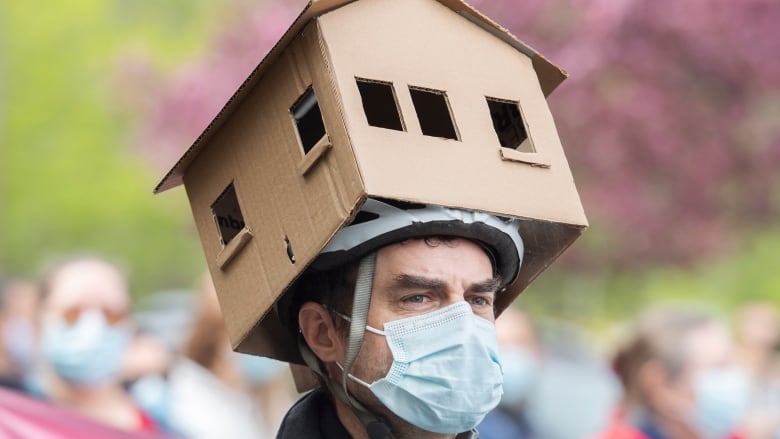 A man with a face mask wears a cardboard house on his head atop a bike helmet. 