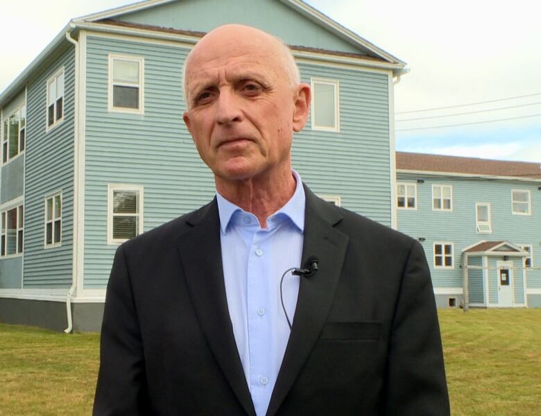 A person wearing a blue shirt and black blazer stands outside on a lawn.