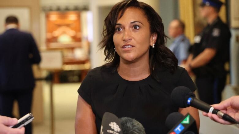 A Black woman wearing a black top is while speaking into reporter's mics