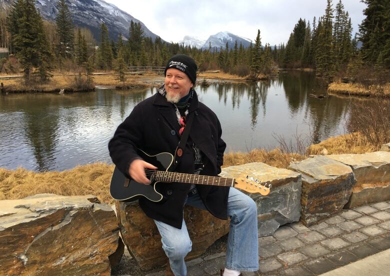 A bearded man holds a guitar while sitting on a stone ledge in front of a body of water and mountains in the distance.