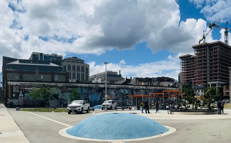 A park on a sunny day, with two white trucks, people standing underneath an orange overhang, with a long, graffiti-ed building in the background. 