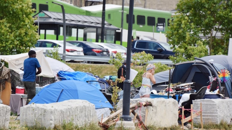 Tents are set up in a vacant lot. Two people can be seen facing away from the camera - a man and a woman. A green and white GO train is seen in the background.