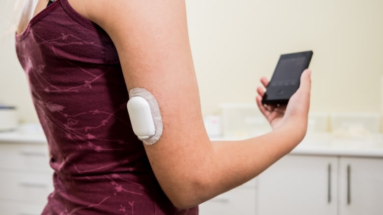 A woman holds a cellphone in her right hand, while a tubeless insulin pump, about three centimetres wide by six centimetres long by one centimetre in thickness, is attached to the back of her upper arm.