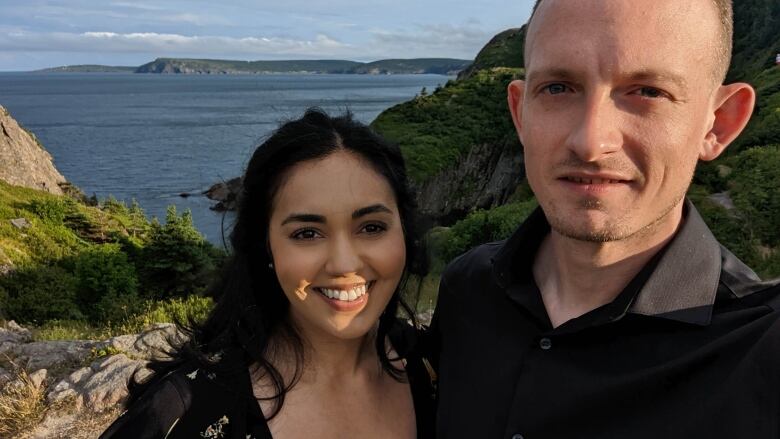 A woman and a man stand in the sun on a hill with the ocean behind them.