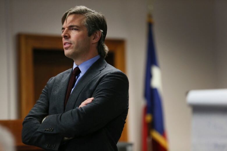 A man in a suit stands in a courtroom with his arms crossed. 