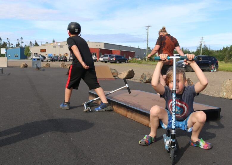 A young child sits on a scooter while holding the handles. Behind, two older children are playing. 