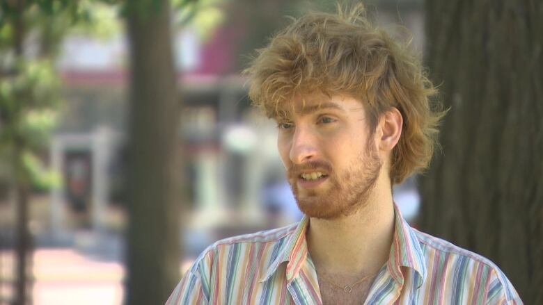 A white man with blonde hair wearing a striped shirt stands outside in front of a tree.