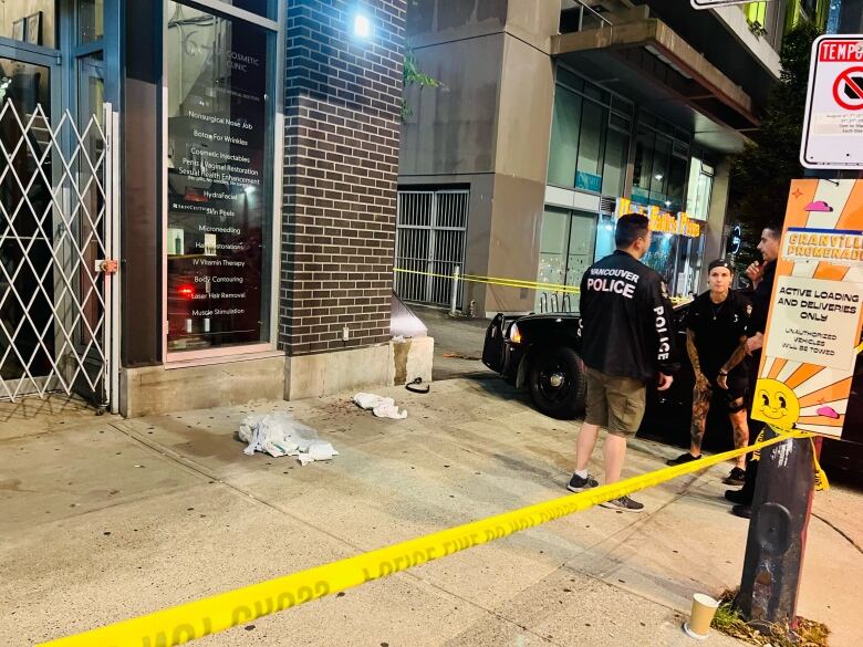 A section of sidewalk is blocked off with yellow caution tape. There are what appear to be cloths and blood stains on the ground and a man with a Vancouver Police Department jacket stands on the corner next to a police car.