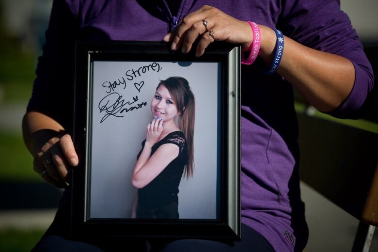 A woman holds a framed photo of a teenaged girl with long brown hair wearing a black dress. The words 