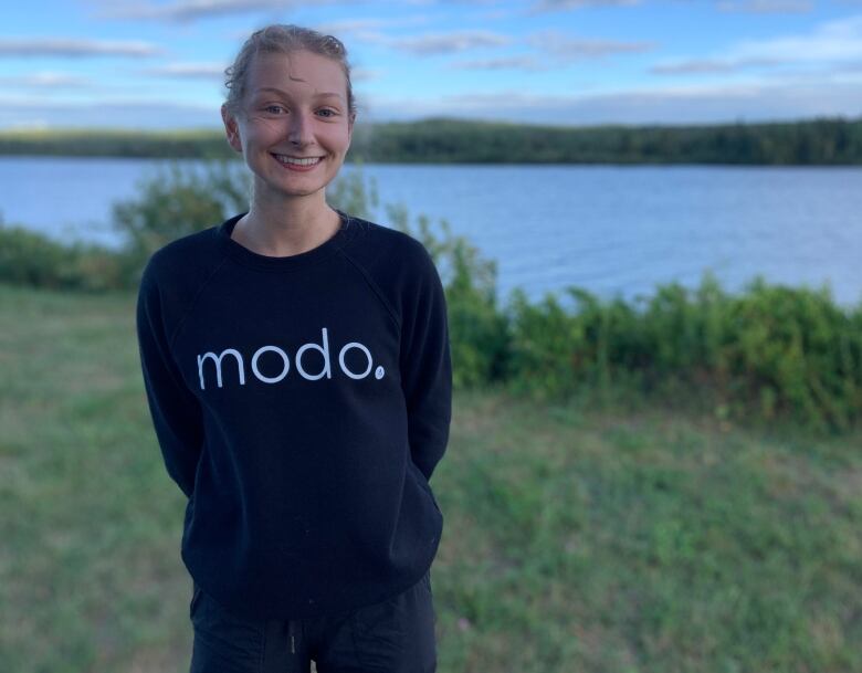 A person stands on a grassy bank in front of a body of water. They're wearing a black sweater with the word 