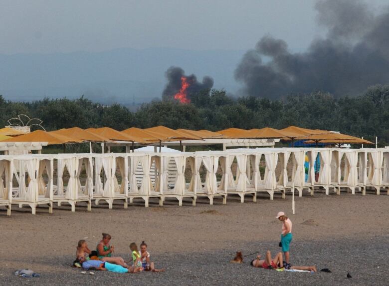 People on a beach. Black smoke and flames seen rising in the background. 