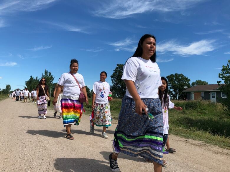 Women and girls of various ages, wearing white T-shirts and ribbon skirts, walk down a gravel road.