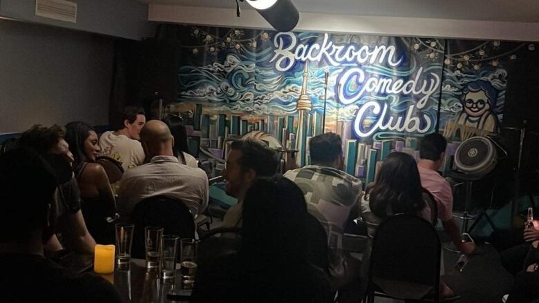 Photo of a seated audience at the Backroom Comedy Club. It is a darkened room packed with people seated at small tables. The stage backdrop is a graffiti-style mural of the Toronto skyline. Type reads: 