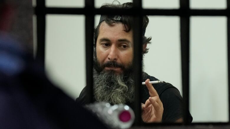 A bearded man holds a cigarette as he speaks. 