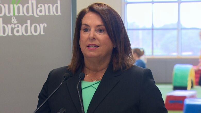 A woman in a black suit stands in front of a sign that says Newfoundland and Labrador sign, with a kids' gymnastics room in the background. 