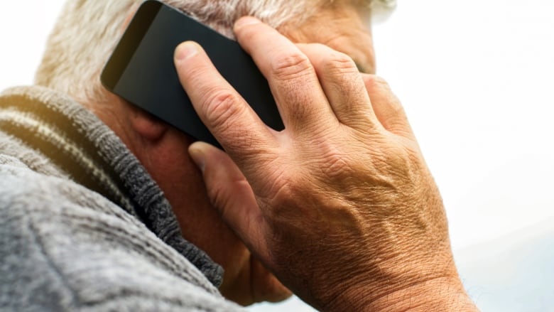 A closeup of a white-haired man in a sweater holding a cellphone up to his ear.