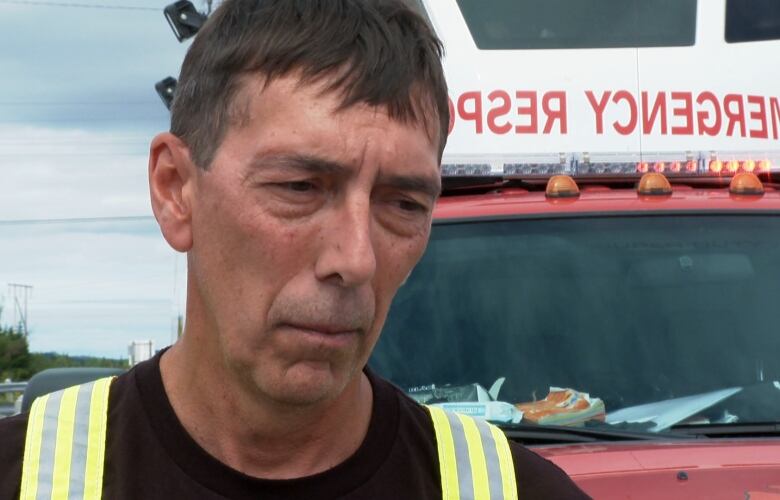 A man in bunker hear stands in front of a fire truck with the emergency lights on. 