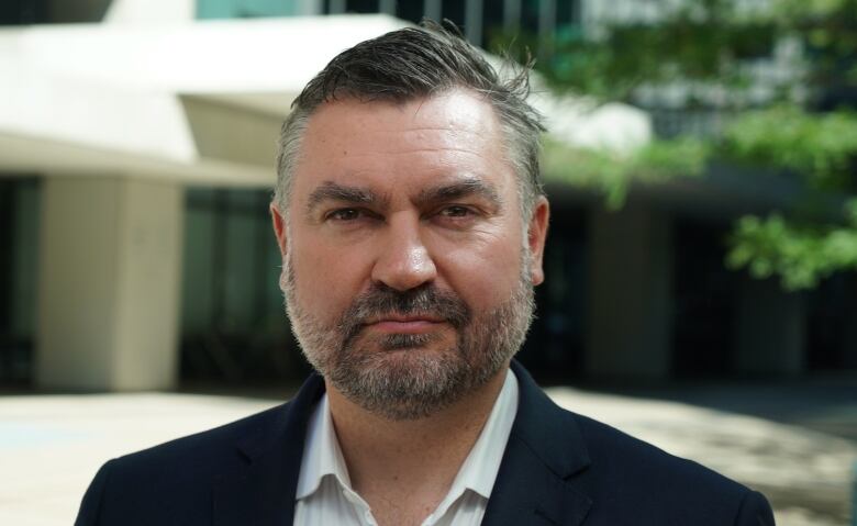 A man with dark hair and stubble is not smiling into the camera.