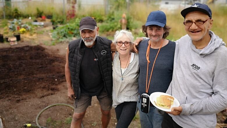 A group of people in a community garden smile. One of them is holding a plate of food.