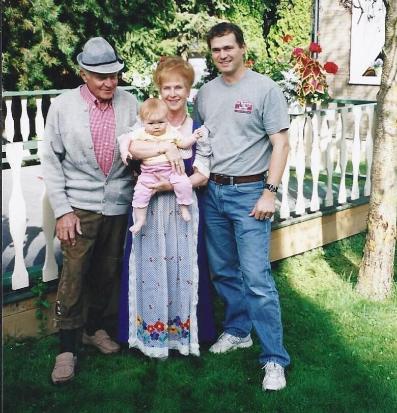 Three people dress up in traditional German attire. The person in the centre, a woman, is holding a baby. The woman is dressed up in a traditional blue dress. The person on the left, a man, is wearing a grey hat and blazer. The man on the right is wearing a grey t-shirt and jeans.