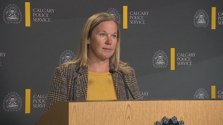 A woman in a yellow shirt and grey jacket stands behind a podium and in front of a backdrop emblazoned with the Calgary Police Service crests. 