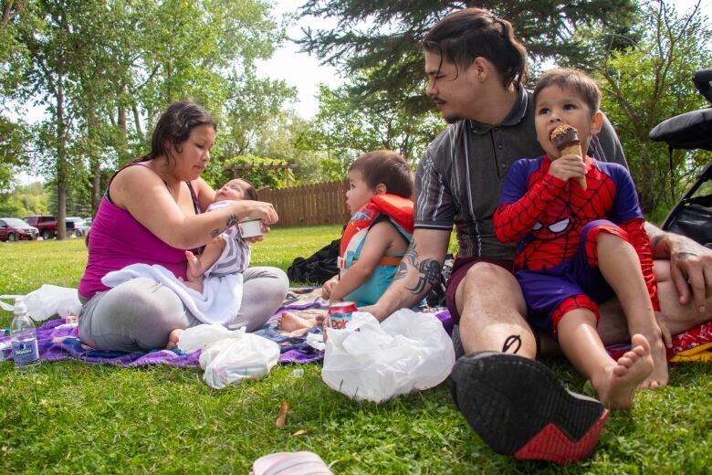 Foreground, a boy in a Spider-Man shirt sits on a man's lap. Behind them are a young boy in a life-jacket and a woman cradling a baby in her arms.