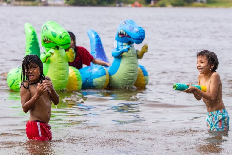 Two young children in swimsuits stand almost waist-deep in the water, with colourful floating dinosaurs behind them.