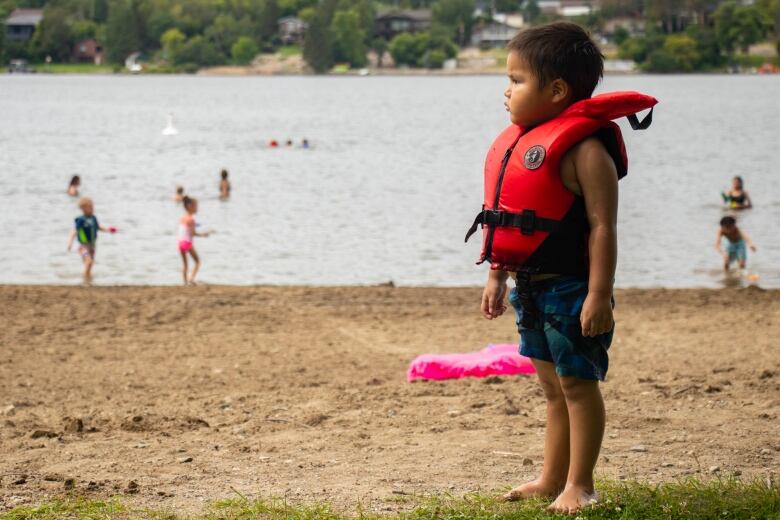 A young boy in an orange life-jacket stands on the beach.