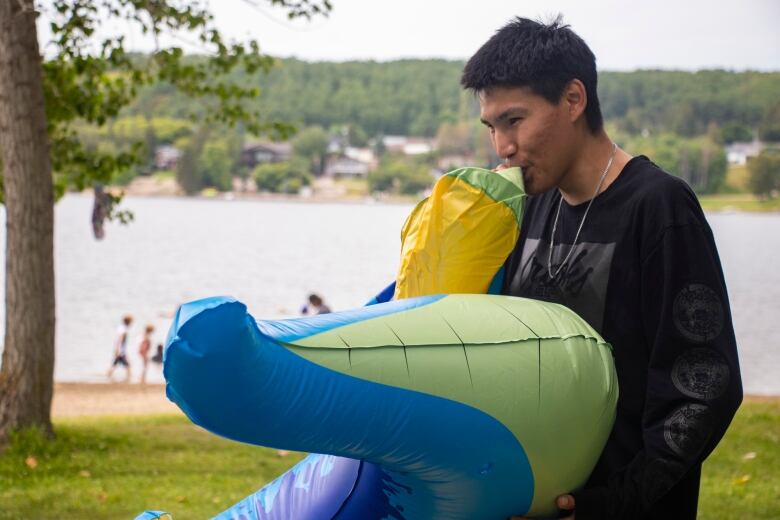 A young man in a black shirt blows air into a blue and green floating plastic dinosaur, with the beach and water behind him.