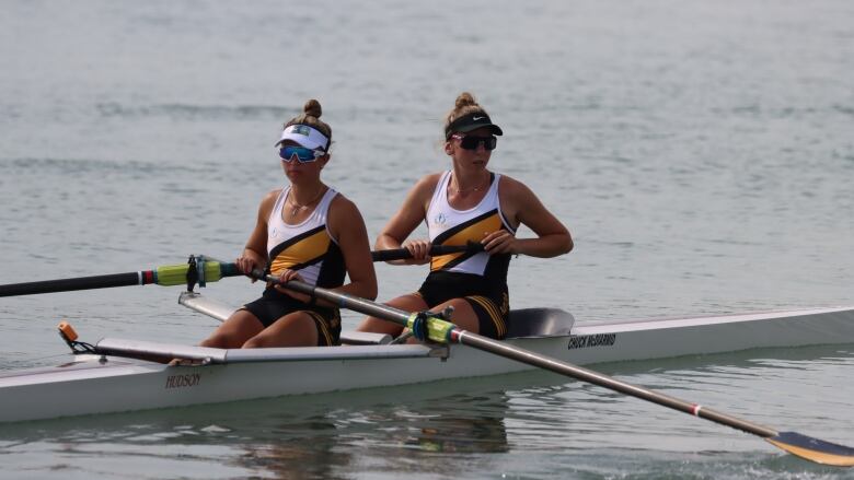 Two female rowers in Manitoba colours, wearing sunglasses and visors, relax in their coxless pair boat at the Canada Summer Games. Leah Miller is in the forward seat, with both hands on her left-side oar, while Katie Sierhuis is in the rear seat, holding the left-side oar.