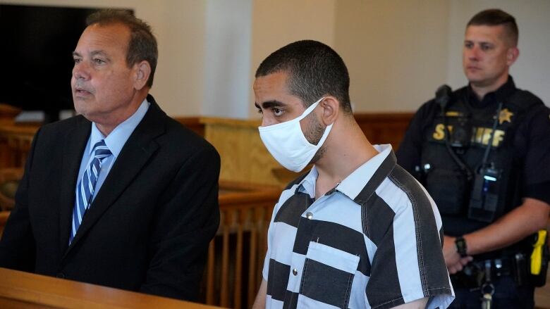 A man in a COVID-19 mask and a prison-issued shirt listens in a court session.