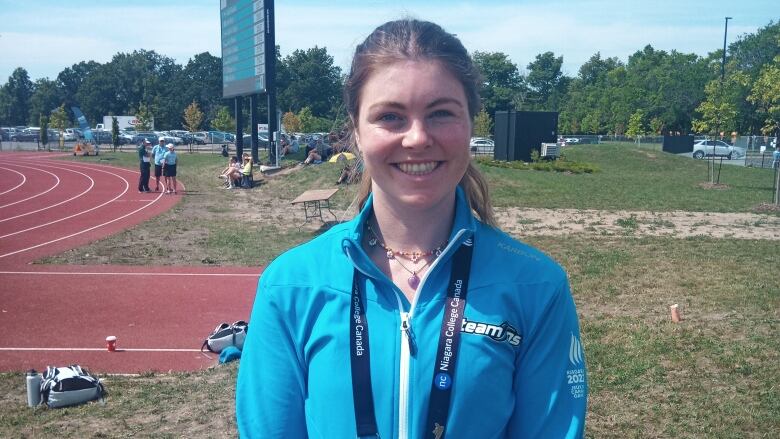 A woman in a blue track jacket stands beside a track with a scoreboard in the background. She is smiling.